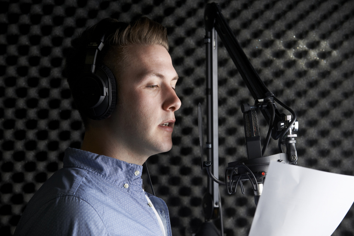 An image featuring a voice-over artist in a recording studio. The artist is positioned in front of a microphone with headphones on, speaking animatedly into the microphone. A soundproofed wall is visible in the background, highlighting the professional setting. A script on a stand is in front of the artist, indicating the ongoing recording session.