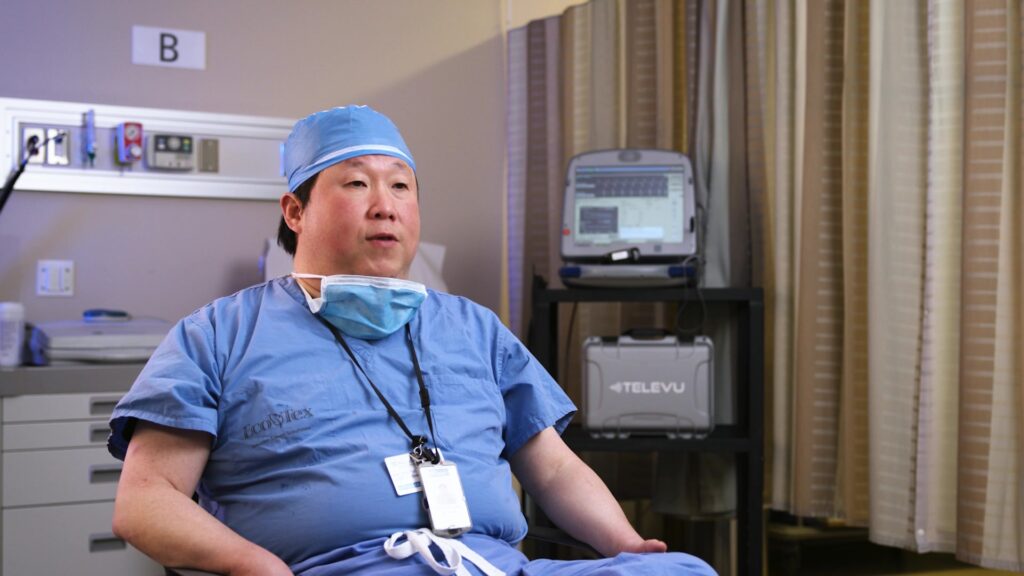 A doctor showing the relaxed pose, leaning casually against a chair during an interview.